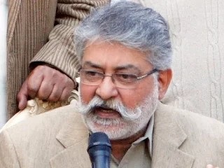 A portrait of Pir Pagara, a bearded man wearing a white turban and a black coat, smiling and looking at the camera.