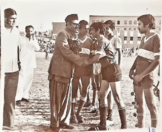 1974 Football Tournament Final at Gangoti Ground, Rohri Black and white photo of Syed Ghulam Shabbir Shah Rizvi MPA, father of Syed Nasir Hussain Shah, shaking hands with a player at the 1974 football tournament final at Gangoti Ground, Rohri, with players and spectators in the background.