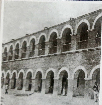 1940s Boarding House A black and white photograph of a two-story building with multiple arches on both levels. The building appears to be made of brick and has a series of evenly spaced arches on the ground floor and the first floor. There are a few people visible near the arches on the ground floor.