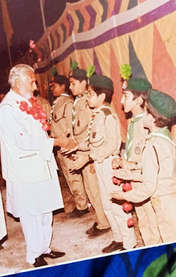 Late Ghulam Siddique Qadri Sahib with Scouts – A Moment of Recognition and Respect Qadri Sahib, adorned with a red flower garland, shaking hands with members of a scout or sports team in blue Adidas uniforms during an outdoor gathering.