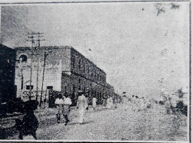 Street Scene Near Sindh Madressa Boarding House, 1940 A black and white photograph of a street scene from 1940, showing people walking along a road with a large building on the left side. The building has multiple stories and features arched windows. There are utility poles and wires visible, and the street appears to be busy with pedestrians.