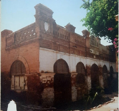 Historic Brick Structure with Arched Windows – Sukkur Heritage Site Old brick building with arched windows and decorative upper facade, partially damaged at the base, with a flowering tree on the right.