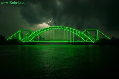 Lansdowne Bridge Illuminated β Independence Day Glow Night view of Lansdowne Bridge in Rohri, Pakistan, illuminated with green LED lights spiraling around its steel arches, reflecting in the Indus River under a dramatic sky.