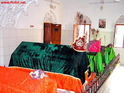 Resting Places of Rizvi Saints – Jan Allah Shah Shrine, Rohri Interior of Jan Allah Shah shrine in Rohri, Sindh, showing graves of Syed Jan Muhammad Shah Rizvi and other Rizvi saints. Covered in green, red, and orange cloths with peacock feathers and decorative crowns.