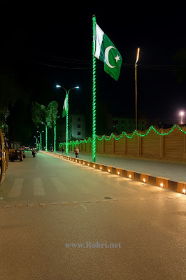 Nighttime Street Decorated for Pakistan Day in Sukkur A nighttime street scene in Sukkur decorated with green LED lights and Pakistani flags. Lampposts are wrapped in glowing green lights, and flags with crescent and star designs line the road. The street is illuminated with festive lighting, showing buildings, a pedestrian crossing, and light traffic, creating a calm and celebratory atmosphere.