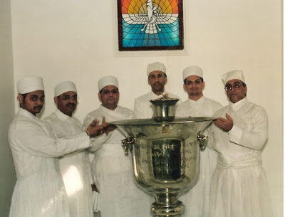 Zoroastrian Ritual Around Inscribed Urn – Faravahar Window & Parsi Khabar 2020 Five individuals in white traditional attire gathered around a large metallic urn with Gujarati inscriptions, beneath a stained glass Faravahar symbol.