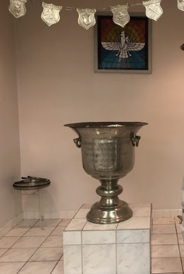 Zoroastrian Ritual Setup with Inscribed Urn and Faravahar – Parsi Khabar 2020 Metallic urn with inscriptions placed on a tiled pedestal, beneath a stained glass Faravahar symbol, with ceremonial plaques and a tray nearby.