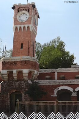 Sukkur Railway Clock Tower: A British Colonial Landmark Near Garam Godi Historic red brick Clock Tower at Sukkur Railway Workshop, Sindh, Pakistan, featuring a large white clock face, arched windows, and colonial architecture against a clear sky with surrounding trees and iron fence.