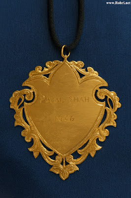 Gold Medal Awarded to Syed Panjal Shah for Rohri Peace Efforts in Manzilgah Riots Close-up view of the gold medal (sono bulo) awarded to Syed Mir Panjal Shah by the Hindu community of Rohri in 1940 for his role in maintaining peace during the Manzilgah riots in Sindh; ornate shield-shaped pendant engraved with 'PANJAL SHAH' and 'NO 4/6', hanging on a black cord against a blue background.