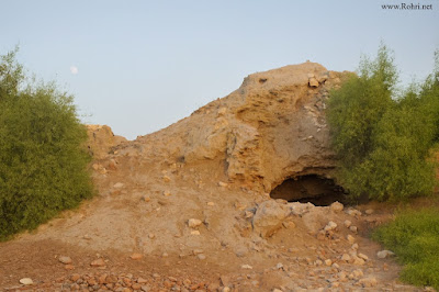 Cave Entrance in Aror (Alor) Ruins Hill β Ancient Sindh Site Natural rock cave entrance in the hills of ancient Aror (Alor) ruins surrounded by green bushes and rocky path under blue sky in Sindh
