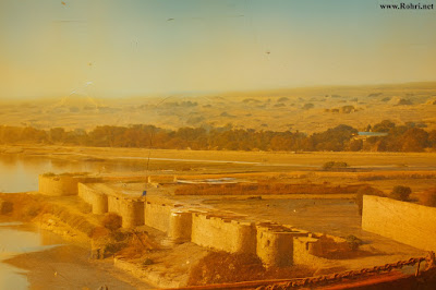 Bukkur Fort – Riverside Bastions Overlooking the Indus Colorized aerial view of Bukkur Fort’s cylindrical bastions along the Indus River in Sindh, Pakistan. The image shows ancient stone walls, a purple flag atop one tower, calm river waters, and a distant townscape with trees and hills—capturing the architectural and cultural heritage of the region.