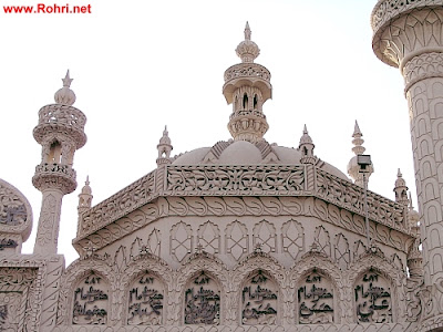 Majestic Facade of Jan Allah Shah Shrine – Arabic Inscriptions and Ornate Sindhi Craftsmanship White stone facade of Jan Allah Shah Janshahi shrine in Rohri, Sindh, featuring Arabic inscriptions, ornate floral carvings, and multiple domes and minarets.