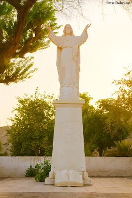 Jesus Christ Statue Sukkur – VENITE AD ME OMNES Inscription and Open Bible Pedestal White marble statue of Jesus Christ with raised arms on a pedestal inscribed with VENITE AD ME OMNES, surrounded by trees and greenery in Sukkur