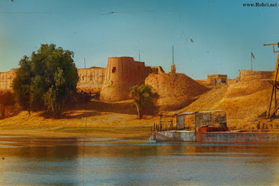 Riverside Fort – Historic Bastion Overlooking the Indus Colorized historical image of a riverside fort with cylindrical mudbrick bastions, palm trees, and a docked barge on the Indus River. The fort stands on a raised bank near Sukkur, Sindh, showcasing early 20th-century architecture and riverine activity.”