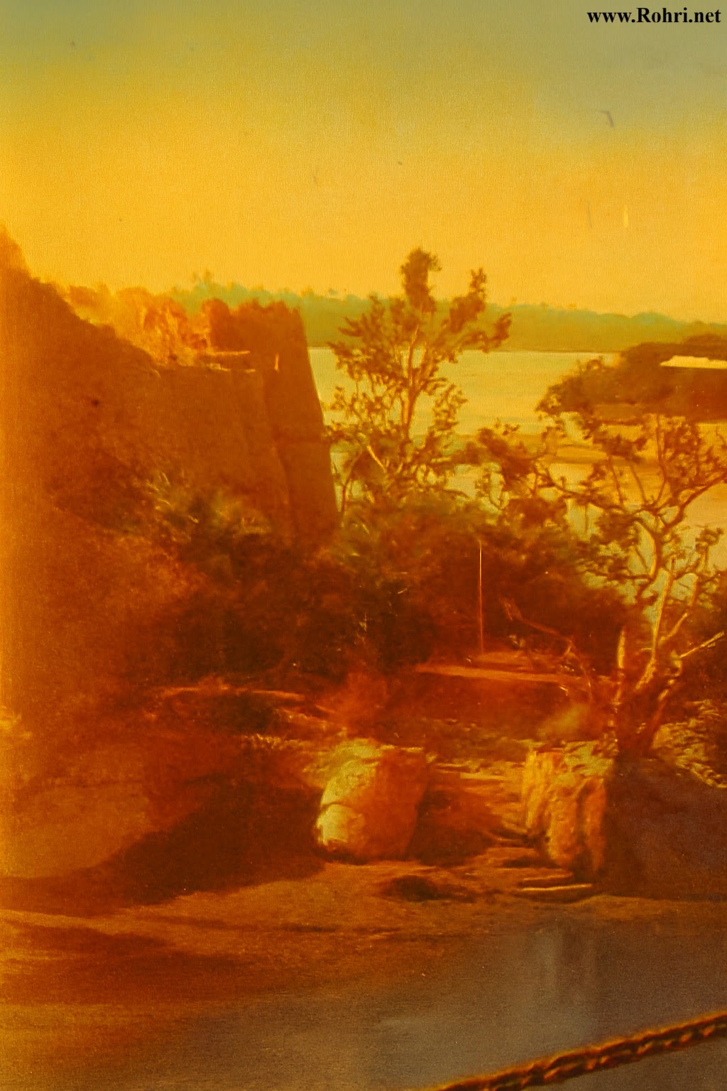 Riverside Bastion – Fortress Wall Overlooking the Indus Colorized historical image of a fortress wall with cylindrical bastion overlooking the Indus River in Sindh, Pakistan. The scene includes rocky terrain, a gnarled tree, palm foliage, and a calm river reflecting the golden hues of sunset—capturing the architectural and natural heritage of the region.