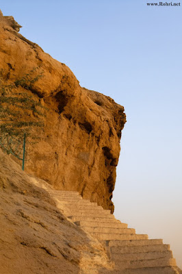 Cliffside Staircase to Shah Maqsood Site – Rohri Heritage Pathway A steep stone staircase carved into a rugged cliff in Rohri, Sindh, leading upward toward Shah Maqsood’s heritage site, with textured rock formations and a sparse tree under a clear sky.