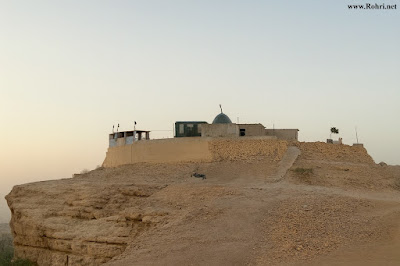 Shah Maqsood Hilltop Shrine – Elevated Heritage Site in Rohri, Sindh A hilltop shrine complex in Rohri, Sindh, featuring a green dome, fortified stone walls, black flags, and a rocky slope with a dirt path leading up, captured under a clear sky at golden hour.