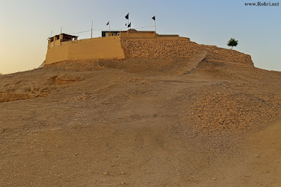 Hilltop Shrine of Shah Maqsood – Sacred Landmark in Rohri, Sindh A fortified hilltop shrine in Rohri, Sindh, with black flags on tall poles, stone walls, and a rugged dirt path leading up the slope, set against a clear sky at dawn or dusk.