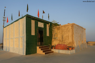 Shah Maqsood Shrine Complex – Rooftop Heritage Site in Rohri, Sindh A rooftop shrine complex in Rohri, Sindh, featuring a green-painted entrance with white lattice panels, colorful flags on the roof, and an adjacent beige structure with a red patterned cloth and potted plants.