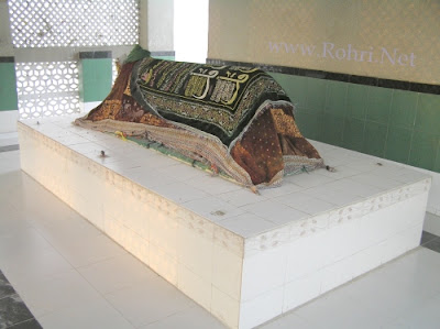 Tomb of Shah Maqsood – Revered Shrine in Rohri, Sindh The tomb of Shah Maqsood in Rohri, Sindh, covered with decorative cloths featuring Islamic calligraphy, set within a tiled mausoleum interior with green walls and a perforated screen.