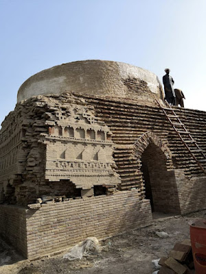Shaikh Laloo Pir Shrine – Restoration of Ancient Brick Dome in Rohri, Sindh Shaikh Laloo Pir Shrine under restoration in Rohri, Sindh, showing scaffolding, a worker near the dome, intricate brickwork with arched niches and geometric patterns, and construction materials like bricks and a red barrel in the foreground.