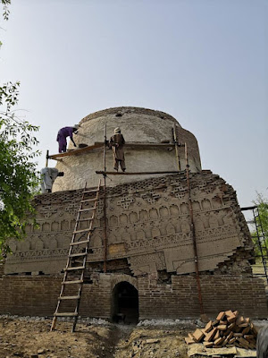 Shaikh Laloo Pir Shrine – Restoration of Ornate Brick Dome in Rohri, Sindh Restoration work on Shaikh Laloo Pir Shrine in Rohri, Sindh, showing scaffolding around an ancient brick structure with a cylindrical dome and intricately carved lower facade featuring arches and geometric patterns, under clear skies.