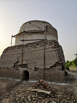 Shaikh Laloo Pir Shrine – Brick Dome Restoration and Ornate Facade in Rohri, Sindh Shaikh Laloo Pir Shrine in Rohri, Sindh, under restoration with scaffolding around a cylindrical brick dome and intricately carved lower facade featuring arched niches and geometric brick patterns, surrounded by debris and trees.